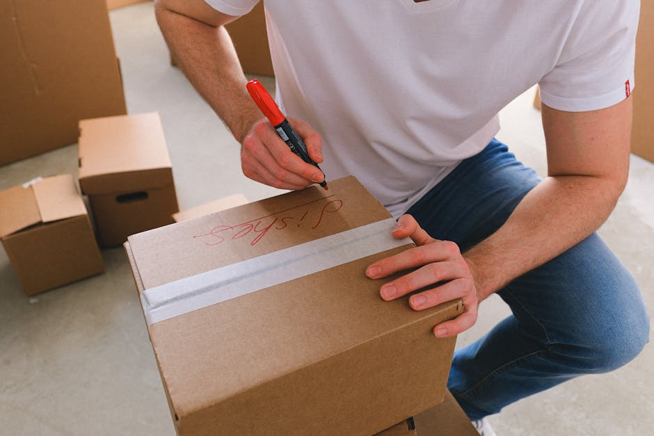 A person wearing a white t-shirt and blue jeans is crouching indoors beside a cardboard box, using a black and red marker to write 'Spares' on the top of the box. Several other cardboard boxes of varied sizes are visible in the background, some sealed with clear packing tape and others open, resting on the floor. The setting appears to be a home or storage area, with light-colored flooring and a partially visible door or wall in the background. The image illustrates packing and organization activities involved in house removals and furniture transport, with the individual preparing boxes for a home relocation. South Lambeth Removals’ services focus on efficient packing, moving logistics, and loading processes to facilitate smooth house removals across Lambeth.