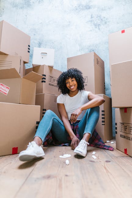 A young woman with curly black hair, wearing a white T-shirt, light blue jeans, and white sneakers, sitting on a wooden floor inside a room surrounded by numerous cardboard moving boxes of various sizes, some sealed with red tape and others with printed labels. She is smiling and looking directly at the camera, with one arm resting on her knee and the other on a box beside her. The boxes are stacked against a light blue textured wall, indicating an active home relocation or packing process. Small packing materials, such as bubble wrap or packing paper, are scattered on the floor near her. The scene suggests a furniture transport or moving preparations, with South Lambeth providing professional removals services for house moves. Overall, the image captures the packing and loading process involved in home relocation, emphasizing the importance of careful packing and organisation during furniture transport.