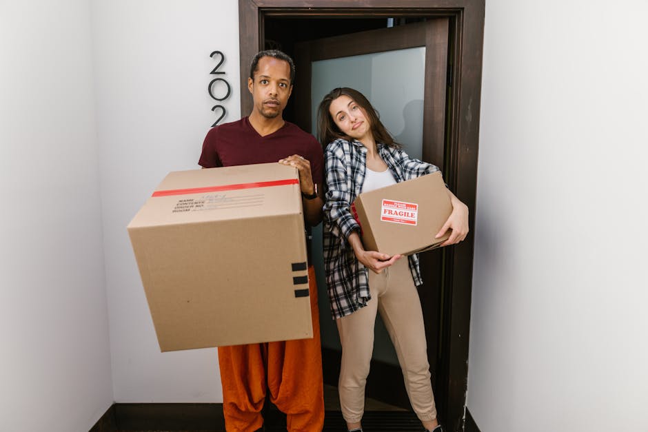 Two women engaged in packing and relocation tasks inside a residential space, surrounded by several large cardboard boxes sealed with red and black packing tape, some of which have shipping labels, indicating a home removal process. The woman on the left, wearing a plaid shirt and beige pants, is holding a smaller box, while the woman on the right, dressed in a white top and patterned trousers, is carrying a large decorative vase made of woven material with a tall green plant. The background features a textured, light-colored wall, and the area is lit by natural or soft artificial light, illustrating a typical packing stage during furniture transport and house removals. This scene reflects professional moving activities managed by South Lambeth, emphasizing packing and loading for a house relocation, with the women actively preparing belongings for transport in a methodical and organized manner.