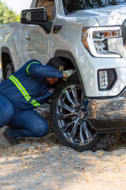 A man wearing a blue uniform with reflective green stripes is kneeling on a gravel surface next to a white pickup truck, inspecting or adjusting the front right wheel. The vehicle is parked near a residential area with trees and foliage in the background. The scene suggests the man is performing a roadside check or maintenance as part of a home relocation or furniture transport process, in an outdoor environment with natural lighting. The image reflects aspects of moving logistics, such as vehicle inspections during transportation, which relates to South Lambeth's removals and moving services for house relocations.