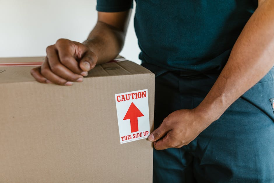 A professional mover from South Lambeth is seen handling a large cardboard box during the home relocation process. The mover's hands are gripping the box, which is sealed with packing tape, and a red and white caution sticker indicating 'This Side Up' with an upward arrow is prominently displayed on the box. The interior setting suggests an entryway or room of a property, with a smooth light-colored wall visible in the background. The mover is dressed casually in a dark t-shirt and dark trousers, and appears to be preparing to load or transport the box onto a van or storage area as part of the furniture transport and packing process. Visible packing materials, such as cardboard and packing tape, highlight the careful handling associated with moving services. South Lambeth's removals team is engaged in the logistics of packing and moving items securely, demonstrating typical procedures involved in professional house removals and ensuring the safe transfer of belongings in a home relocation scenario.