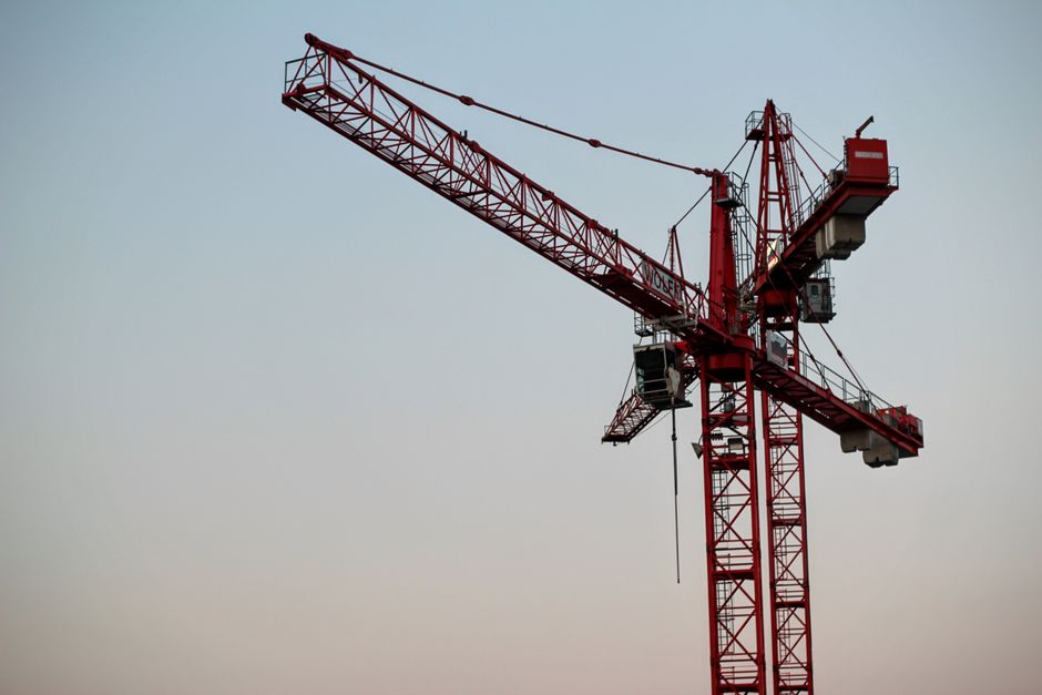 A high-rise construction site featuring a tall modern glass building under construction, with a green construction hoist labeled 'Spring' attached to the exterior for vertical transport of workers and
