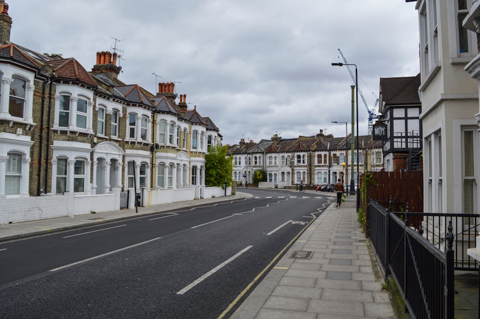 The image depicts a residential street scene with row houses featuring white facades, large bay windows, and tiled roofs, typical of Victorian architecture in South Lambeth. The street is paved with asphalt, with visible lane markings, and has a concrete sidewalk with metal railings along the right side. In the foreground, there are no vehicles or furniture visible on the street, but in the background, a construction crane can be seen indicating ongoing development. The sky is overcast with grey clouds, and street lamps are present along the pavement. The scene is calm, with a person walking along the sidewalk, and the environment appears quiet and orderly, suitable for context of home relocation or furniture transport associated with South Lambeth removals projects.