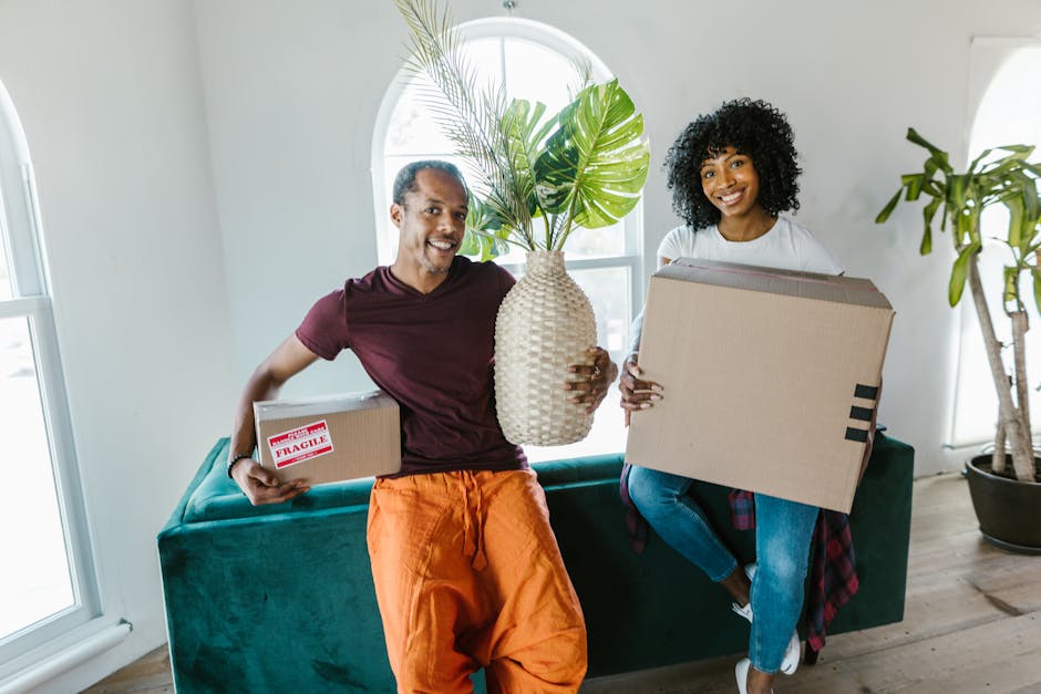 A man and a woman inside a bright, modern living room are holding various items in preparation for a house move. The man, wearing a maroon T-shirt and orange trousers, is holding a small cardboard box with a 'Fragile' sticker, while the woman, dressed in a white T-shirt and jeans, is carrying a larger cardboard box. Both are smiling and looking at the camera. They are positioned near a green sofa with a large potted plant and a window behind them, allowing natural light to illuminate the space. The scene reflects an organized home relocation process, with packing supplies visible, inside a well-lit room with wooden flooring. South Lambeth removals company occasionally featured, emphasizing professional furniture transport and packing during house moves.