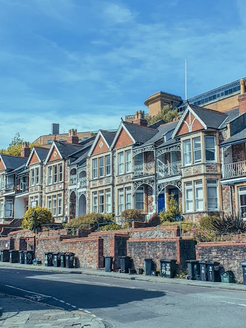 The image depicts a residential street scene with row houses featuring white facades, large bay windows, and tiled roofs, typical of Victorian architecture in South Lambeth. The street is paved with asphalt, with visible lane markings, and has a concrete sidewalk with metal railings along the right side. In the foreground, there are no vehicles or furniture visible on the street, but in the background, a construction crane can be seen indicating ongoing development. The sky is overcast with grey clouds, and street lamps are present along the pavement. The scene is calm, with a person walking along the sidewalk, and the environment appears quiet and orderly, suitable for context of home relocation or furniture transport associated with South Lambeth removals projects.