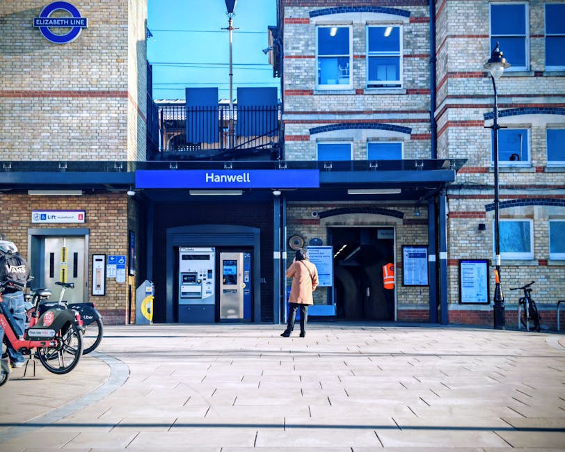 The entrance of Hanwell station, part of the Elizabeth Line, showing a brick-built building with large windows and an elevator door on the left side; a person dressed in a beige coat and black pants stands in front of the station, possibly waiting to load or unload belongings during a home relocation process. Several bicycles are parked nearby, including a bike-sharing scheme with a red Voi electric scooter and a black bicycle secured by a lock on the right. The sidewalk consists of light-colored paving stones, and there are informational displays and signage related to station access and services mounted on the station facade. The scene is well-lit with natural daylight, and the station’s signage indicates it is part of a busy urban transport hub, supporting the logistics involved in furniture transport, packing, or moving operations frequently managed by companies like South Lambeth during house removals or relocations.