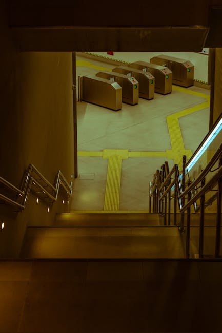 A view looking down from a staircase inside a building towards an underground station entrance with automatic ticket barriers in the background. The staircase has metallic railings on both sides, and the steps are made of dark material. The station floor is illuminated with bright, even lighting, revealing a tiled surface with yellow tactile paving strips guiding towards the barriers. The barriers are rectangular with card scanners on top, positioned in a row across the entrance area. The environment appears clean and well-maintained, suited for facilitating home relocation or moving services in the Vauxhall area, with the station access guide likely indicating the proximity to transport links for furniture transport and packing and moving activities. South Lambeth's removals services would coordinate with such access points for efficient home relocations near Vauxhall station.